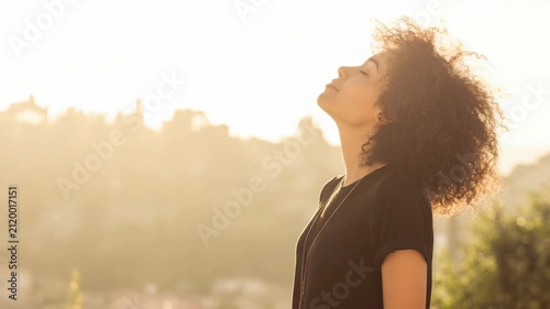Young woman with curly hair breathing fresh air outdoors at sunset, peaceful girl enjoying warm golden hour light in a beautiful scenic landscape