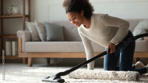 Happy young woman cleaning carpet with vacuum cleaner in living room, smiling African American lady doing housework chores at home, video clip
