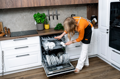 Quadro su tela Young woman homemaker opening dishwasher with clean white dishes modern kitchen