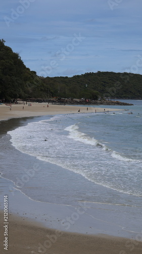 The image depicts a peaceful beach scene with gentle waves approaching the shoreline. The sandy beach is sparsely populated with a few people walking and relaxing along the water's edge