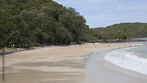 The image depicts a tranquil beach scene with a wide stretch of sandy shoreline bordered by a lush, green hillside on one side