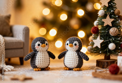 Two cute handmade crocheted amigurumi toy penguins standing together in a beautiful cozy festive miniature room setting with a small sofa, decorated holiday tree and warm bokeh lights.