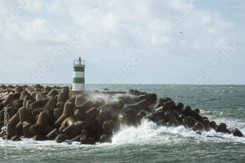 Green and white lighthouse standing on rocky harbor barrier as Atlantic Ocean waves crash along the coast in Portugal beneath bright cloudy sky and dramatic seaside scenery.
