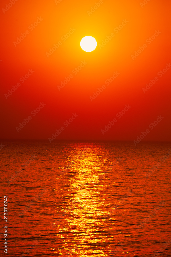 Fototapeta premium Intense vibrant red and orange sunset over quiet sea horizon with bright sun reflection on water.