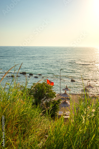Sticker Scenic view of the sea from a grassy cliff with a red Albanian flag and straw beach umbrellas at sunset