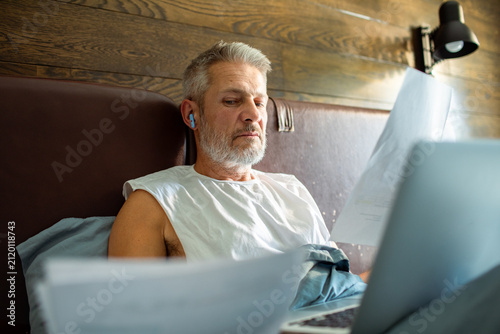Middle aged man reviewing bills on laptop in bed at home