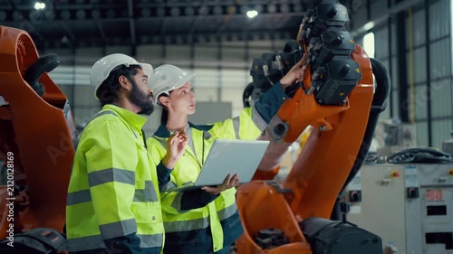 Two engineers stand beside an orange robotic arm. They wear hard hats and high-visibility jackets. One holds a laptop, pointing at the robot