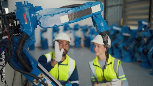Two engineers inspect robotic arm in factory. They wear hard hats and safety vests. They kneel beside blue industrial machinery, discussing operations.