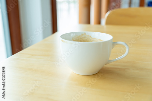 Single, half-empty white ceramic coffee cup with traces of foam and milk stain on the inside rim, sitting on a natural wood grain table in a cafe or breakroom setting.