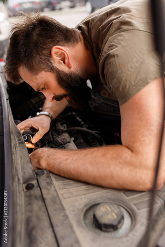 Vertical orientation bearded man mechanic repairing broken car engine under open hood on street auto service inspection close up driver fixing vehicle motor breakdown vehicle troubleshooting maintenan