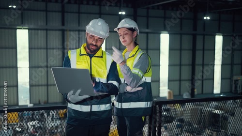 Two engineers review data on a laptop. They stand on a metal platform overlooking a factory floor. Wearing helmets and safety vests