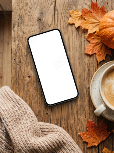 Autumn smartphone mockup with blank screen on a wooden table surrounded by fall leaves, pumpkin, coffee cup and cozy sweater from top view with copy space