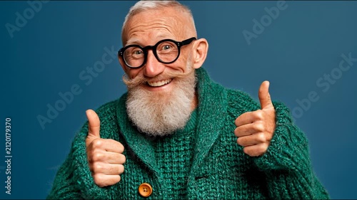 Cheerful senior man with glasses and beard gives double thumbs up against blue background.