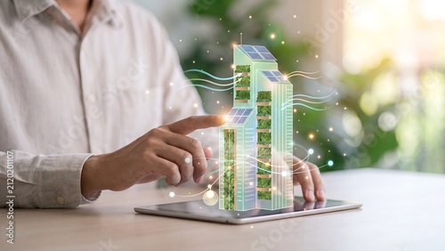 Man pointing at a holographic green building model with solar panels and vegetation above a tablet, representing sustainable architecture, smart city development, and eco-friendly real estate innovati