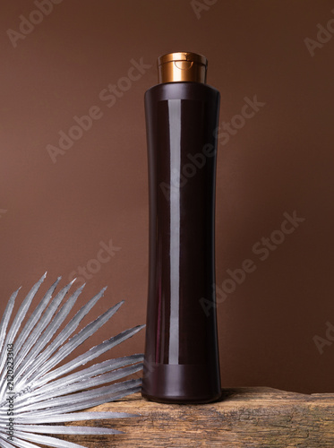A plastic brown cosmetic bottle with a golden flip-top lid stands on a wooden surface. A silver palm branch. Brown background.cosmetic product mockup