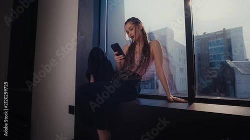 Young woman using smartphone while sitting on city windowsill beside bright urban window glow inside