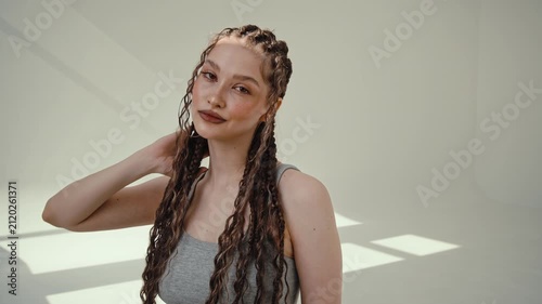 Confident young woman with braided hair and hoop earrings indoors in soft daylight against bare wall
