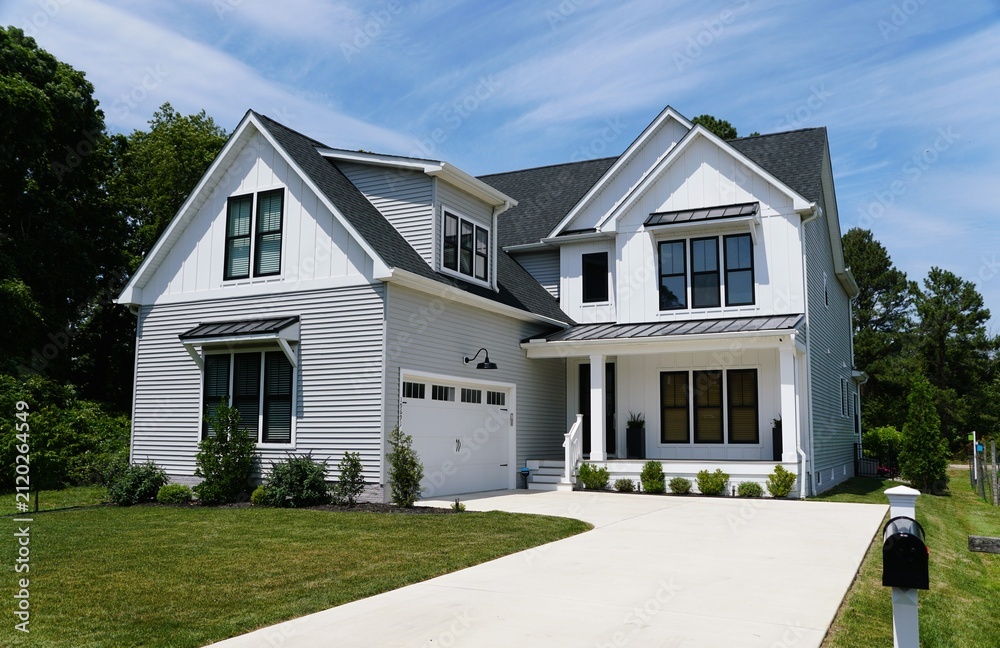 Fototapeta premium A two-story modern suburban home with white siding, black trim, gabled roofs, landscaped yard, and paved driveway leading to double garage near Rehoboth Beach, Delaware, U.S.A