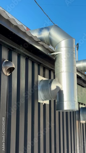 Metal ventilation ducts on corrugated wall under clear blue sky, outdoor exhaust pipes mounted to workshop exterior, steel elbow
