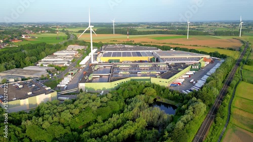 Aerial drone footage of the Colruyt distribution center in Halle, Belgium, showcasing sustainable energy with wind turbines and solar panels at sunset.