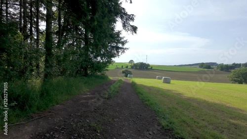 A slow tracking shot moving forward on a dirt path through a peaceful countryside landscape with green fields, a forest, and hay bales on a sunny summer day.