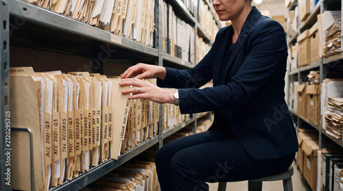 A professional businesswoman searching through hanging file folders on a metal shelf in an office archive room.