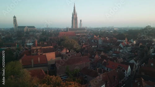 Wide aerial panorama of Bruges, Belgium, featuring Gothic church towers, medieval architecture and dense red rooftops across the historic city at dawn.