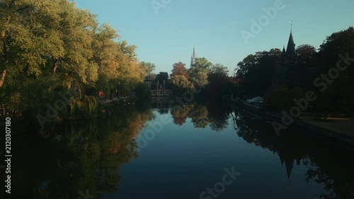 Calm water in Bruges, Belgium, mirrors autumn trees and distant historic buildings, forming a tranquil park landscape beneath clear warm morning light at sunrise.