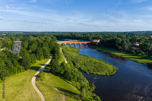 Fotografía Aerial view shows the red brick Kuldiga Bridge over the Venta River in Kuldiga, Latvia, with the broad low Venta Rapid upstream, summer midday light, trees, and town