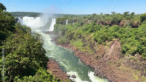 Panoramic view of Iguazu Falls cascading into the river canyon surrounded by dense tropical rainforest under clear blue sky