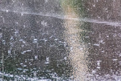 Heavy rain pours down on a dark asphalt surface, creating numerous splashes, ripples, and a shimmering wet texture as raindrops impact the flooded pavement during a powerful downpour.