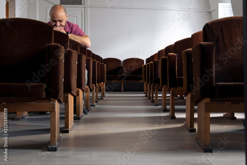 Man praying alone in empty church with rows of velvet chairs