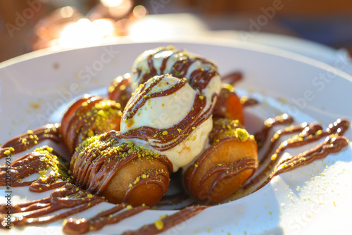 A Greek dessert of golden pastries topped with ice cream, chocolate drizzle, and pistachios in Santorini, Greece, glowing in warm Mediterranean light.
