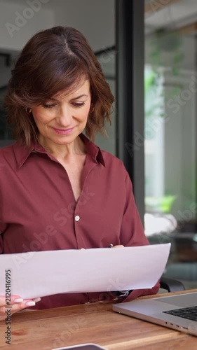 Latin businesswoman checking paper report at desk in office. Mature woman financial analyst holds document, works on laptop. Female expert reviews business contract at workplace. Vertical
