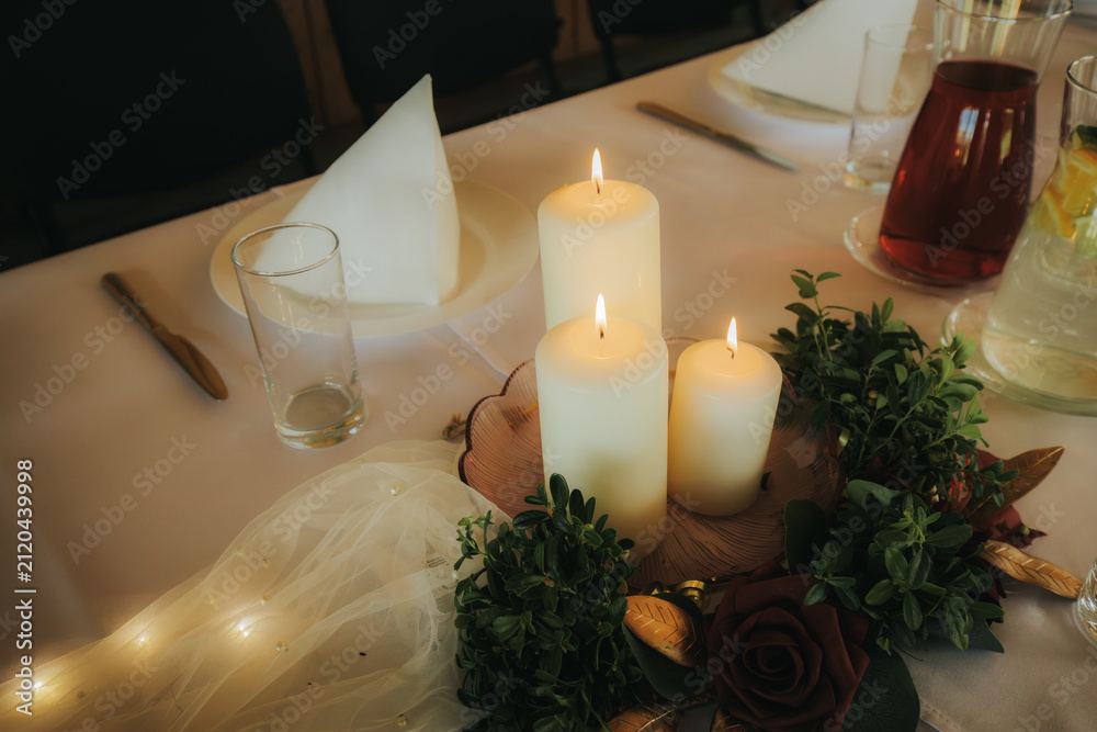 Obraz premium Table setting with three lit candles surrounded by greenery and a decorative rose, elegant dining atmosphere with glassware and cutlery visible in the background