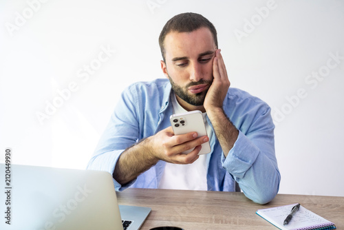 Canvas Print Bored man holding a smartphone, resting his chin on his hand while sitting at a
