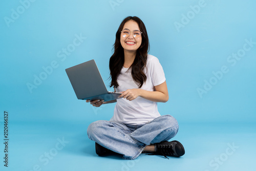Young woman happily working on a laptop while sitting cross-legged on a blue background