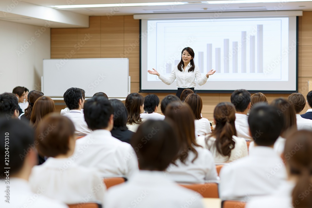 Obraz premium プレゼンテーションを行う日本人女性と真剣に学ぶ聴衆のセミナー風景