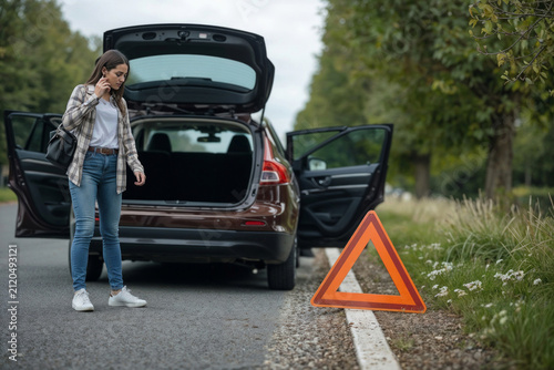 Woman stands beside broken car, distressed, phone to ear. Trunk open, doors ajar, hazard sign placed beside road. Trees line asphalt, wildflowers peek through roadside grass