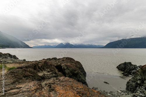 Rocky shoreline overlooking silty water and snow-capped mountains under overcast sky at Beluga Point Turnagain Arm Anchorage Alaska USA