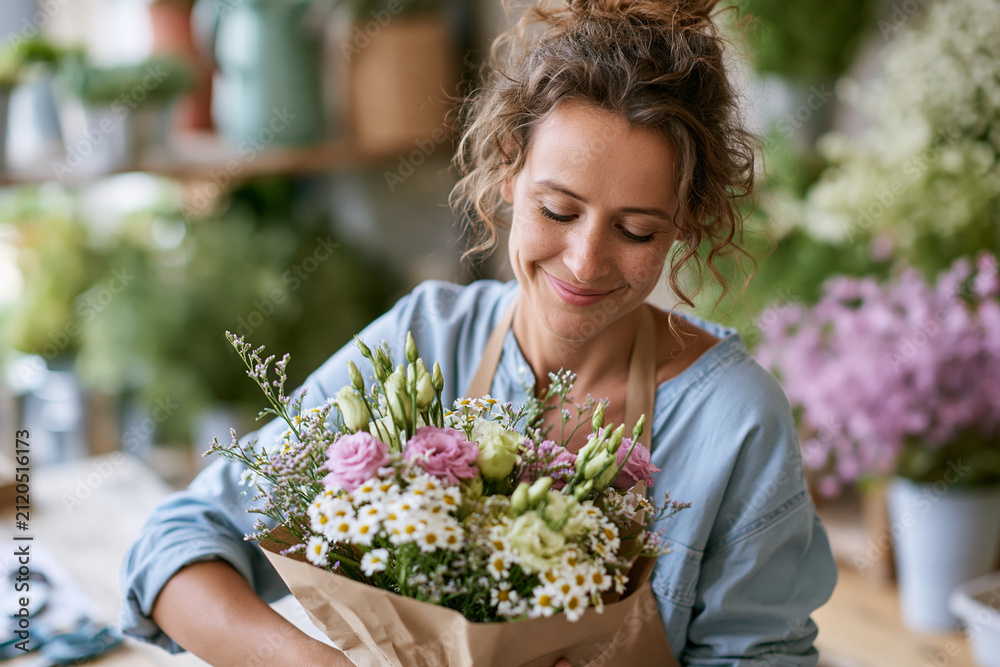 Obraz premium Smiling florist holds a vibrant bouquet wrapped in brown paper. Her curly hair frames her face as she looks down. Soft lighting highlights the delicate pink roses and white daisies