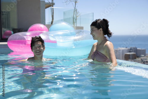 Asian couple laughing and swimming in rooftop infinity pool near floats, woman in striped bikini