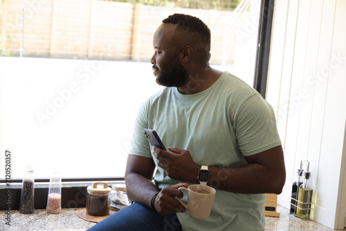 Mid-adult African man sitting on granite countertop by bright window, holding mug and smartphone