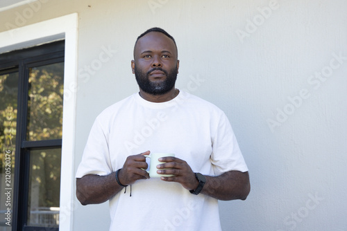 Mid-adult African American man standing at sliding glass door holding white mug and wearing watch