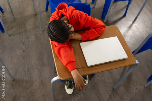 African male teen slumping over school desk in classroom wearing red hoodie, closed white laptop