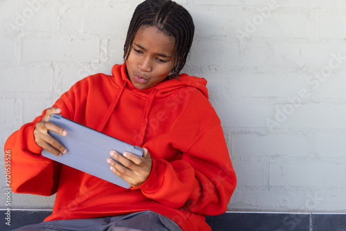 African American teenager in red hoodie sitting against gray wall on dark ledge holding gray tablet