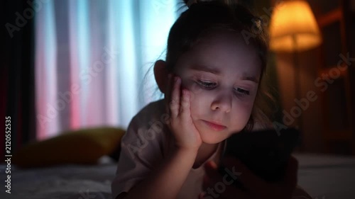 Close-up of a young girl face lying on a bed in the evening, holding a smartphone.
