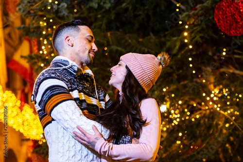 Obraz na plátně Young couple hugging and smiling near a decorated christmas tree at night