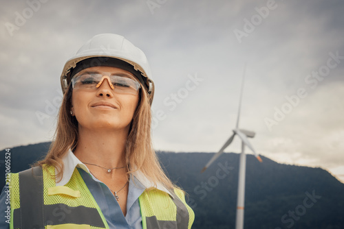 A confident and skilled female engineer, adorned in safety gear, stands proudly at a vast wind farm, showcasing the remarkable dedication and innovative spirit of the sustainable energy sector