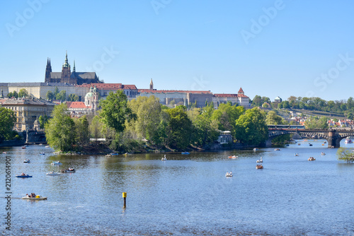 A panoramic view of Prague from the Vltava River, featuring Prague Castle and St. Vitus Cathedral above the historic city, with Charles Bridge and boats visible in the foreground